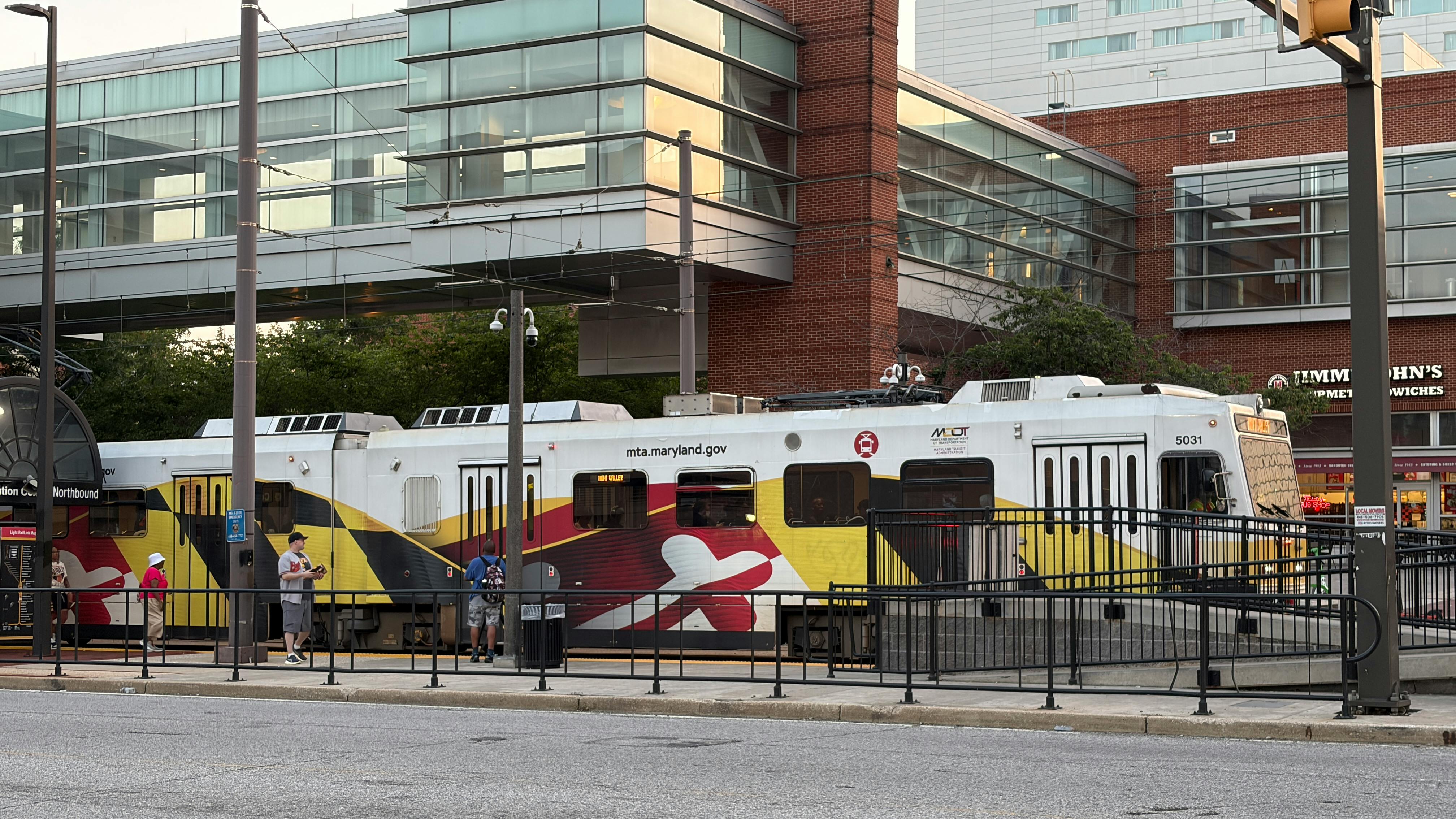 Maryland Transit Administration light-rail car number 5031 in the agency's Maryland state flag-inspired yellow, red, black and white livery stops at a downtown Baltimore station platform bound for Hunt Valley, with riders waiting alongside the train and mixed-use commercial buildings visible in the background.
