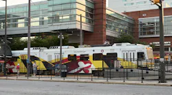 Maryland Transit Administration light-rail car number 5031 in the agency's Maryland state flag-inspired yellow, red, black and white livery stops at a downtown Baltimore station platform bound for Hunt Valley, with riders waiting alongside the train and mixed-use commercial buildings visible in the background.