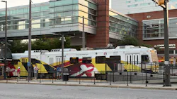 Maryland Transit Administration light-rail car number 5031 in the agency's Maryland state flag-inspired yellow, red, black and white livery stops at a downtown Baltimore station platform bound for Hunt Valley, with riders waiting alongside the train and mixed-use commercial buildings visible in the background.