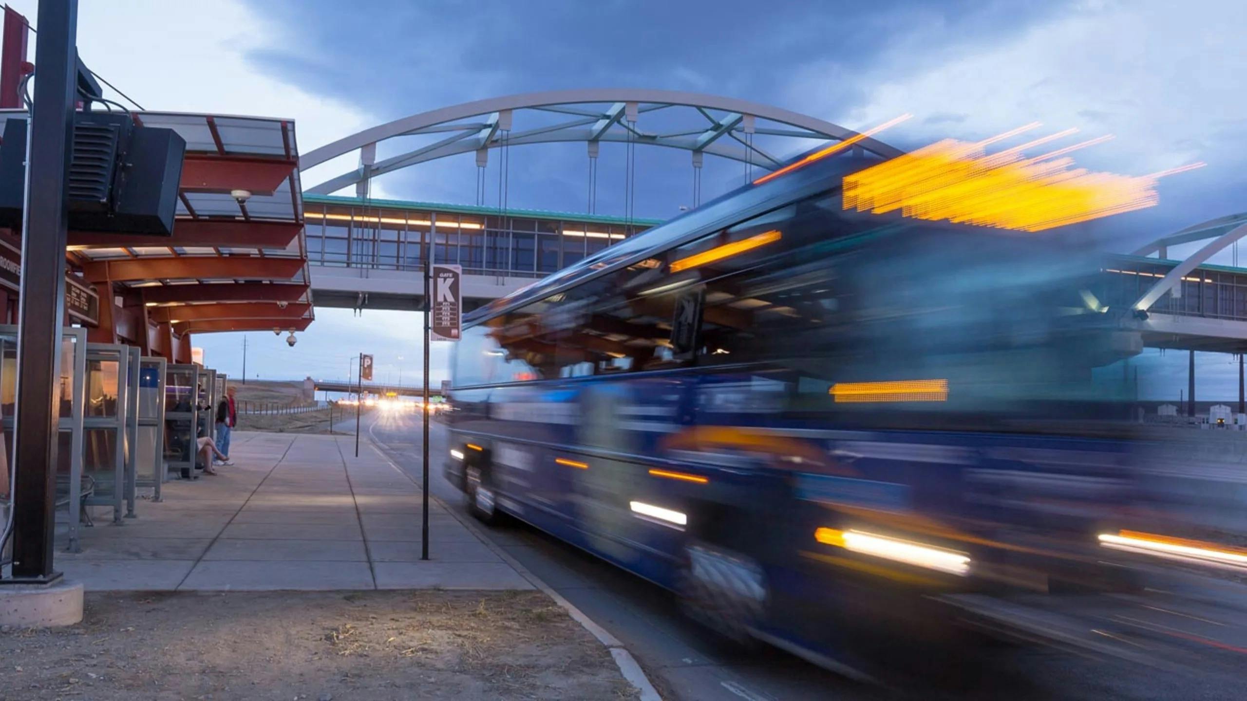 Long-exposure evening photo of an RTD Flatiron Flyer bus rapid transit vehicle passing a station platform in motion blur, with waiting passengers under a red canopy shelter and an arched pedestrian bridge overhead.