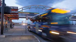 Long-exposure evening photo of an RTD Flatiron Flyer bus rapid transit vehicle passing a station platform in motion blur, with waiting passengers under a red canopy shelter and an arched pedestrian bridge overhead.