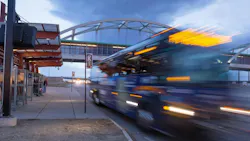 Long-exposure evening photo of an RTD Flatiron Flyer bus rapid transit vehicle passing a station platform in motion blur, with waiting passengers under a red canopy shelter and an arched pedestrian bridge overhead.