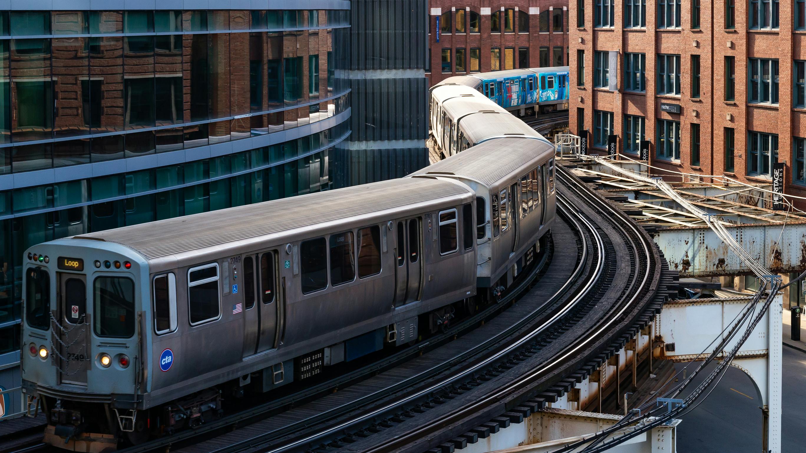 Aerial view of two CTA 'L' trains, including one signed 'Loop,' curving along elevated tracks through downtown Chicago, framed by modern glass towers and historic brick buildings.