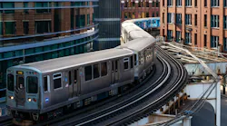 Aerial view of two CTA 'L' trains, including one signed 'Loop,' curving along elevated tracks through downtown Chicago, framed by modern glass towers and historic brick buildings.