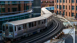 Aerial view of two CTA 'L' trains, including one signed 'Loop,' curving along elevated tracks through downtown Chicago, framed by modern glass towers and historic brick buildings.