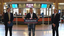 Three officials stand at a press conference inside a historic train station concourse, with the center speaker at a podium bearing a 'Golden Age of Rail — Delivered by President Trump and Secretary Duffy' sign, flanked by two colleagues, with ticket counter screens and the station's Art Deco interior visible in the background.