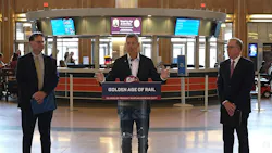 Three officials stand at a press conference inside a historic train station concourse, with the center speaker at a podium bearing a 'Golden Age of Rail — Delivered by President Trump and Secretary Duffy' sign, flanked by two colleagues, with ticket counter screens and the station's Art Deco interior visible in the background.
