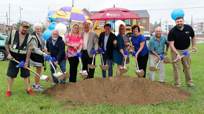 A groundbreaking ceremony with nine officials and community members standing behind a mound of dirt, holding ceremonial shovels tied with blue ribbons, with colorful umbrellas, balloons, and construction equipment in the background.