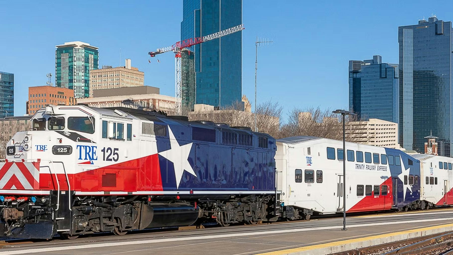 Trinity Railway Express locomotive number 125 in a bold Texas flag-inspired red, white and blue livery with a lone star graphic leads a consist of TRE bi-level passenger cars at a Fort Worth station platform, with the downtown Fort Worth skyline and construction cranes visible in the background under a clear blue sky.