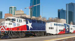 Trinity Railway Express locomotive number 125 in a bold Texas flag-inspired red, white and blue livery with a lone star graphic leads a consist of TRE bi-level passenger cars at a Fort Worth station platform, with the downtown Fort Worth skyline and construction cranes visible in the background under a clear blue sky.