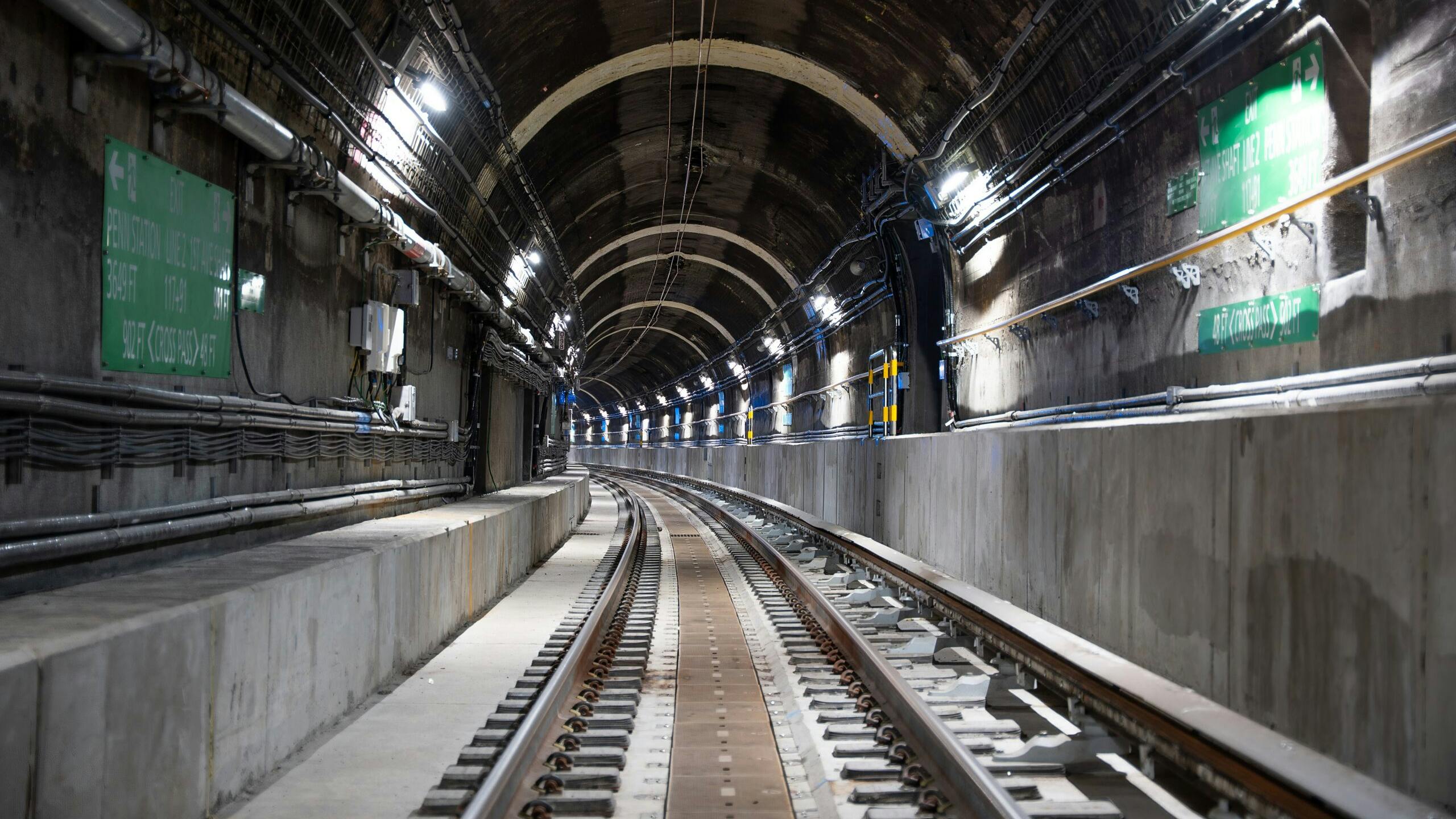 A perspective view down a dark, arched subway tunnel interior showing two sets of tracks curving into the distance, with green wayfinding signage, conduit bundles, utility lines, and overhead lighting mounted along the brick and concrete tunnel walls