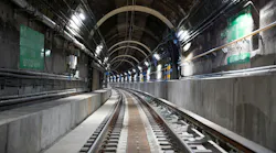 A perspective view down a dark, arched subway tunnel interior showing two sets of tracks curving into the distance, with green wayfinding signage, conduit bundles, utility lines, and overhead lighting mounted along the brick and concrete tunnel walls