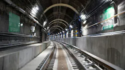 A perspective view down a dark, arched subway tunnel interior showing two sets of tracks curving into the distance, with green wayfinding signage, conduit bundles, utility lines, and overhead lighting mounted along the brick and concrete tunnel walls