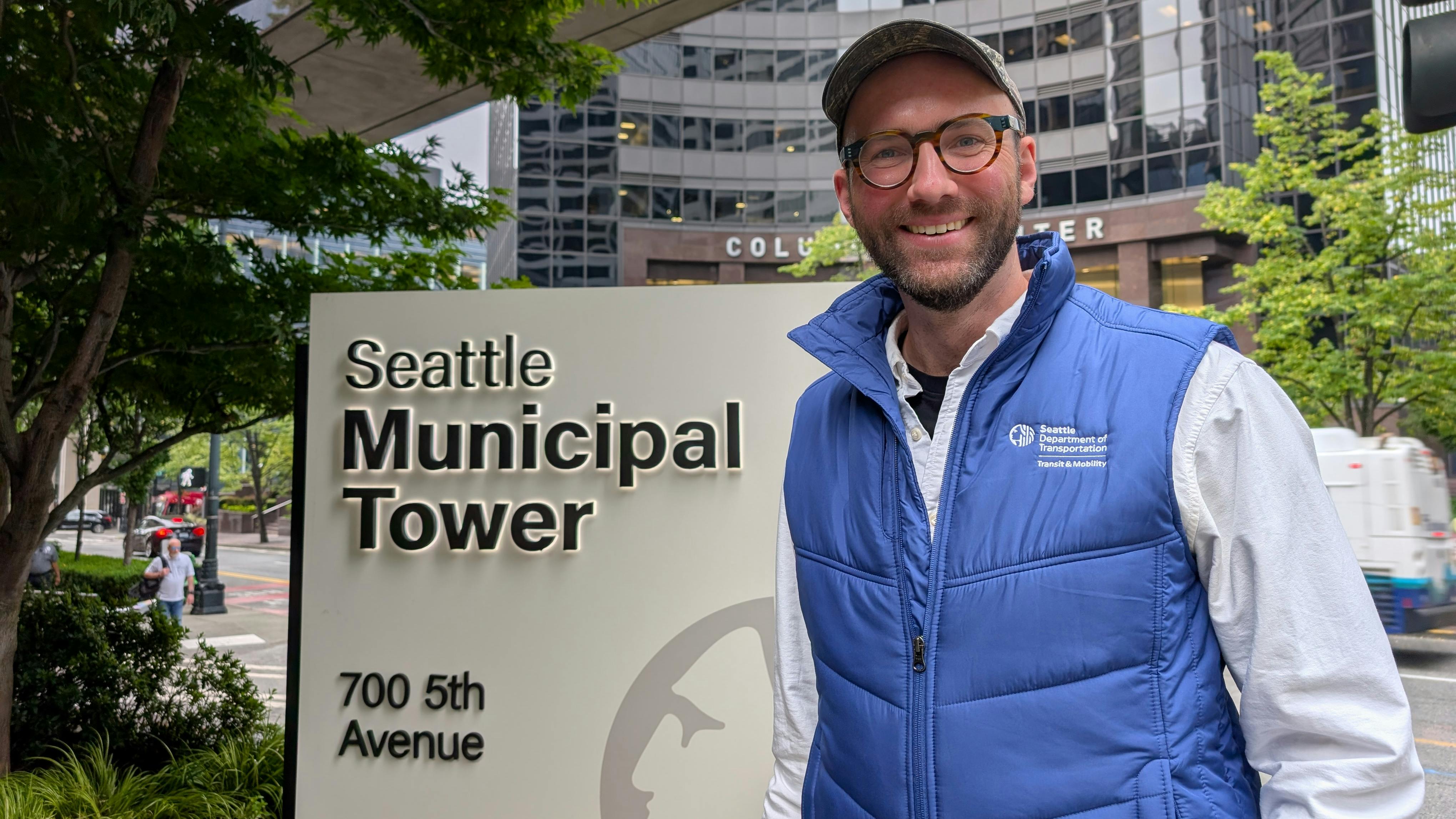Matthew Trecha, wearing a Seattle Department of Transportation Transit & Mobility vest, smiles while standing beside the Seattle Municipal Tower sign at 700 5th Avenue, with the Columbia Center visible in the background.