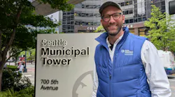 Matthew Trecha, wearing a Seattle Department of Transportation Transit & Mobility vest, smiles while standing beside the Seattle Municipal Tower sign at 700 5th Avenue, with the Columbia Center visible in the background.