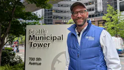 Matthew Trecha, wearing a Seattle Department of Transportation Transit & Mobility vest, smiles while standing beside the Seattle Municipal Tower sign at 700 5th Avenue, with the Columbia Center visible in the background.