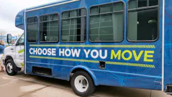 A small transit cutaway bus wrapped in a blue graphic vinyl with the message 'Choose How You Move' in large white and yellow-green lettering, with an All Access Pass branding partially visible near the front door, parked at an outdoor event.
