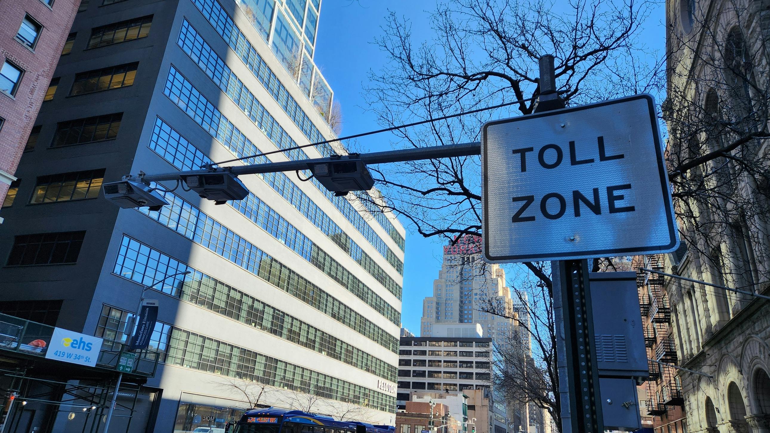 A congestion pricing 'Toll Zone' sign and overhead camera array on a Manhattan street, with an MTA bus and traffic passing below and the New Yorker Hotel sign visible in the distance.