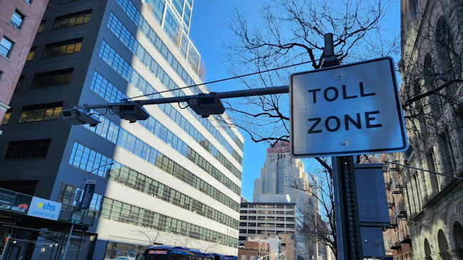 A congestion pricing 'Toll Zone' sign and overhead camera array on a Manhattan street, with an MTA bus and traffic passing below and the New Yorker Hotel sign visible in the distance.