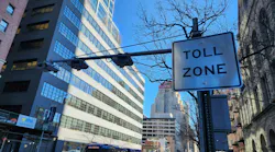 A congestion pricing 'Toll Zone' sign and overhead camera array on a Manhattan street, with an MTA bus and traffic passing below and the New Yorker Hotel sign visible in the distance.