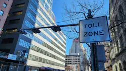A congestion pricing 'Toll Zone' sign and overhead camera array on a Manhattan street, with an MTA bus and traffic passing below and the New Yorker Hotel sign visible in the distance.