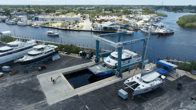Aerial view of the San Francisco Bay Ferry vessel 'Bay Breeze' being hoisted out of the water by a Marine Travelift boat lift at Tarpon Marine Services, surrounded by other boats in dry dock and a waterfront marina in the background.