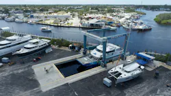 Aerial view of the San Francisco Bay Ferry vessel 'Bay Breeze' being hoisted out of the water by a Marine Travelift boat lift at Tarpon Marine Services, surrounded by other boats in dry dock and a waterfront marina in the background.