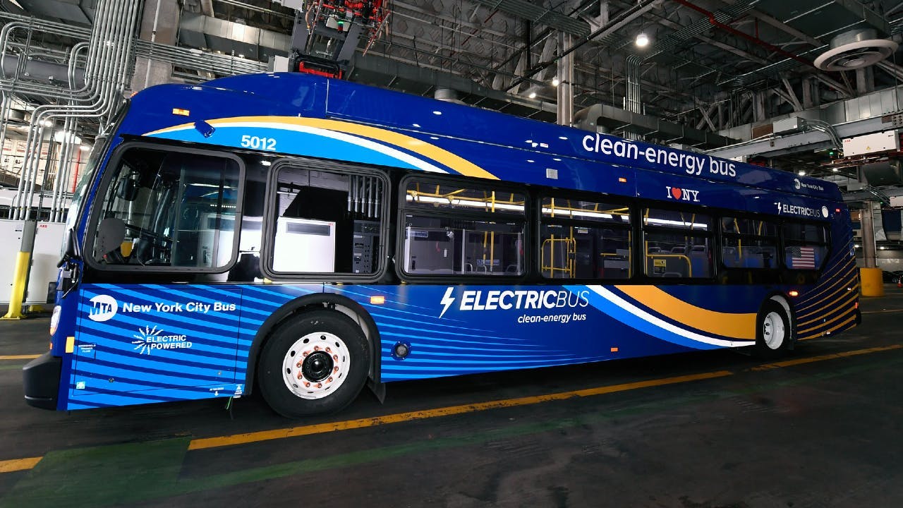 MTA New York City Bus electric bus number 5012 in a deep blue and gold wave livery with 'Electric Bus &mdash; Clean-Energy Bus' and 'I ❤ NY' branding displayed inside a bus depot, with charging infrastructure and industrial ceiling systems visible in the background.