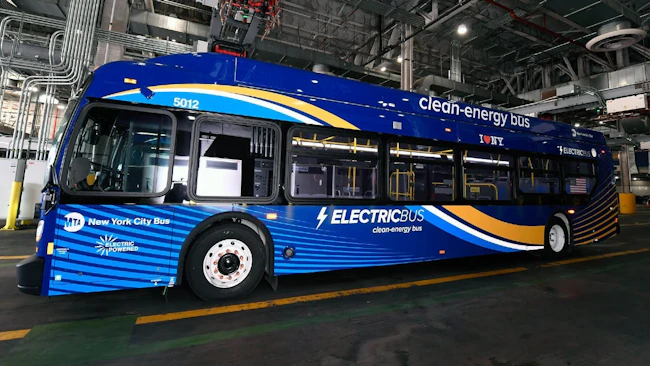 MTA New York City Bus electric bus number 5012 in a deep blue and gold wave livery with 'Electric Bus &mdash; Clean-Energy Bus' and 'I ❤ NY' branding displayed inside a bus depot, with charging infrastructure and industrial ceiling systems visible in the background.