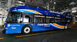 MTA New York City Bus electric bus number 5012 in a deep blue and gold wave livery with 'Electric Bus — Clean-Energy Bus' and 'I ❤ NY' branding displayed inside a bus depot, with charging infrastructure and industrial ceiling systems visible in the background.