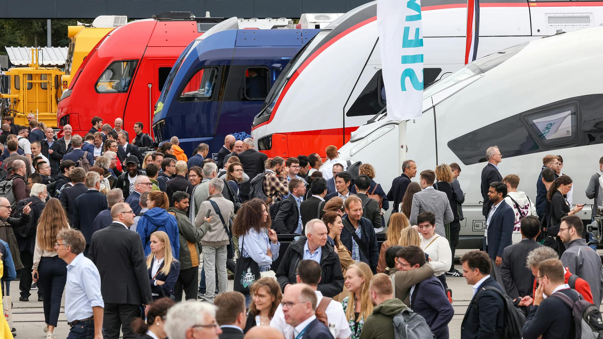 people walk past light rail vehicles at the outdoor display