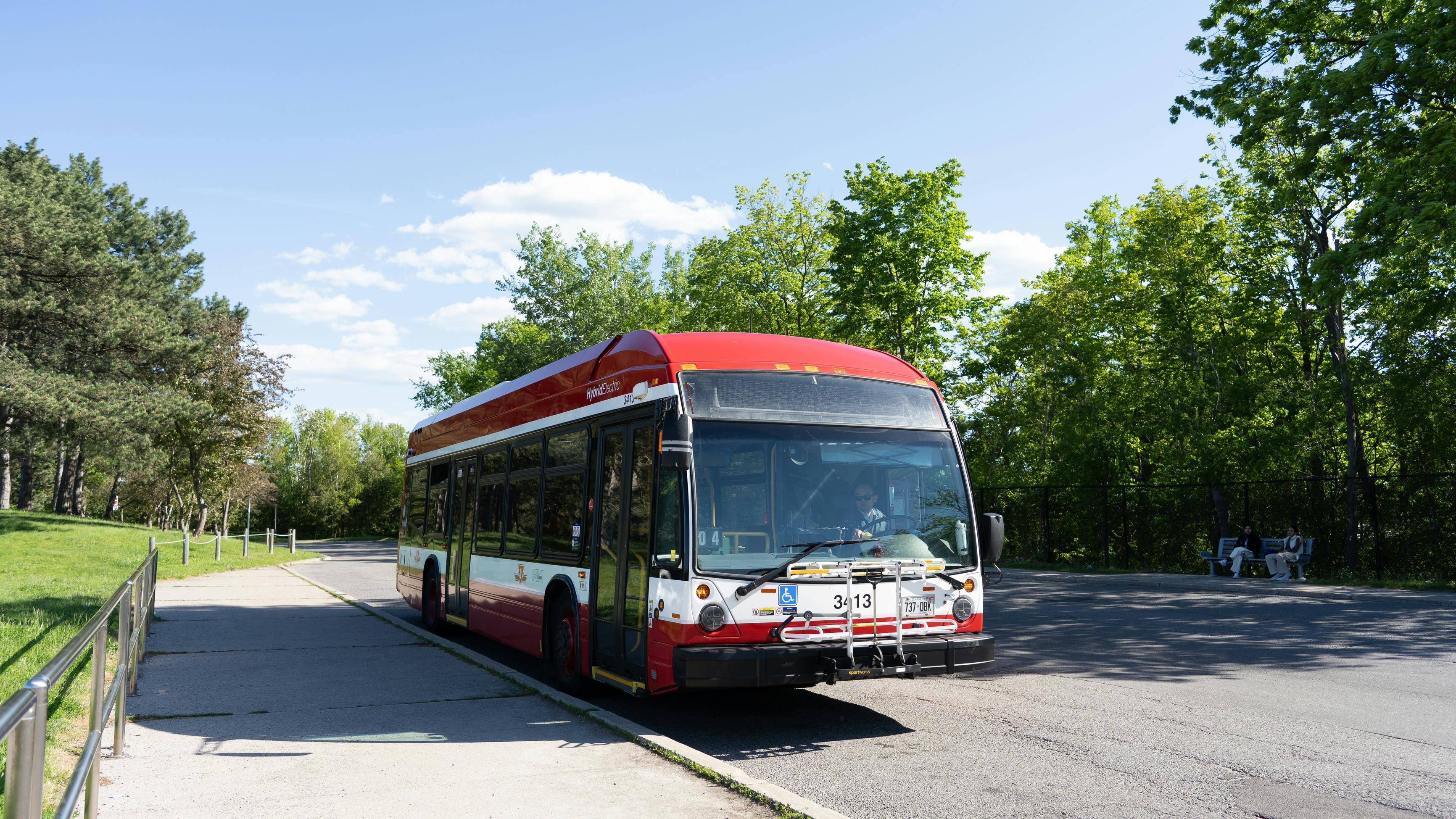 A TTC hybrid-electric bus, numbered 3413, approaching a park-adjacent stop on a paved path, with a front bike rack and accessibility symbol visible, and passengers seated on a nearby bench under trees.