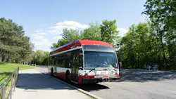 A TTC hybrid-electric bus, numbered 3413, approaching a park-adjacent stop on a paved path, with a front bike rack and accessibility symbol visible, and passengers seated on a nearby bench under trees.