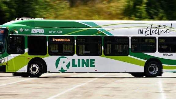 RIPTA R Line electric bus number 2201 in a green and white livery with 'I'm Electric!' branding passes by in motion, displaying 'Training Bus' on its destination board.