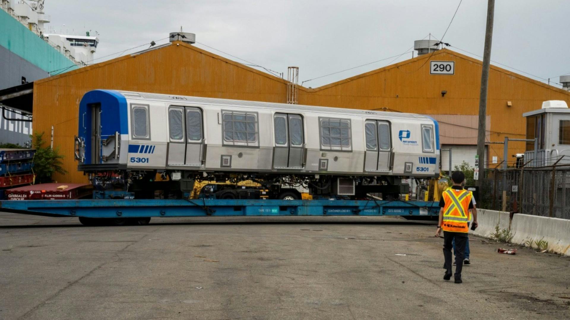 A new Port Authority Trans-Hudson (PATH) rail car, numbered 5301, sits on a flatbed trailer near a shipping port, with a worker in a safety vest walking toward it and orange warehouse buildings in the background.