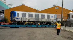 A new Port Authority Trans-Hudson (PATH) rail car, numbered 5301, sits on a flatbed trailer near a shipping port, with a worker in a safety vest walking toward it and orange warehouse buildings in the background.