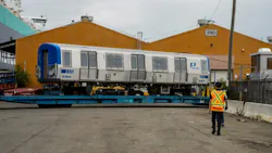A new Port Authority Trans-Hudson (PATH) rail car, numbered 5301, sits on a flatbed trailer near a shipping port, with a worker in a safety vest walking toward it and orange warehouse buildings in the background.