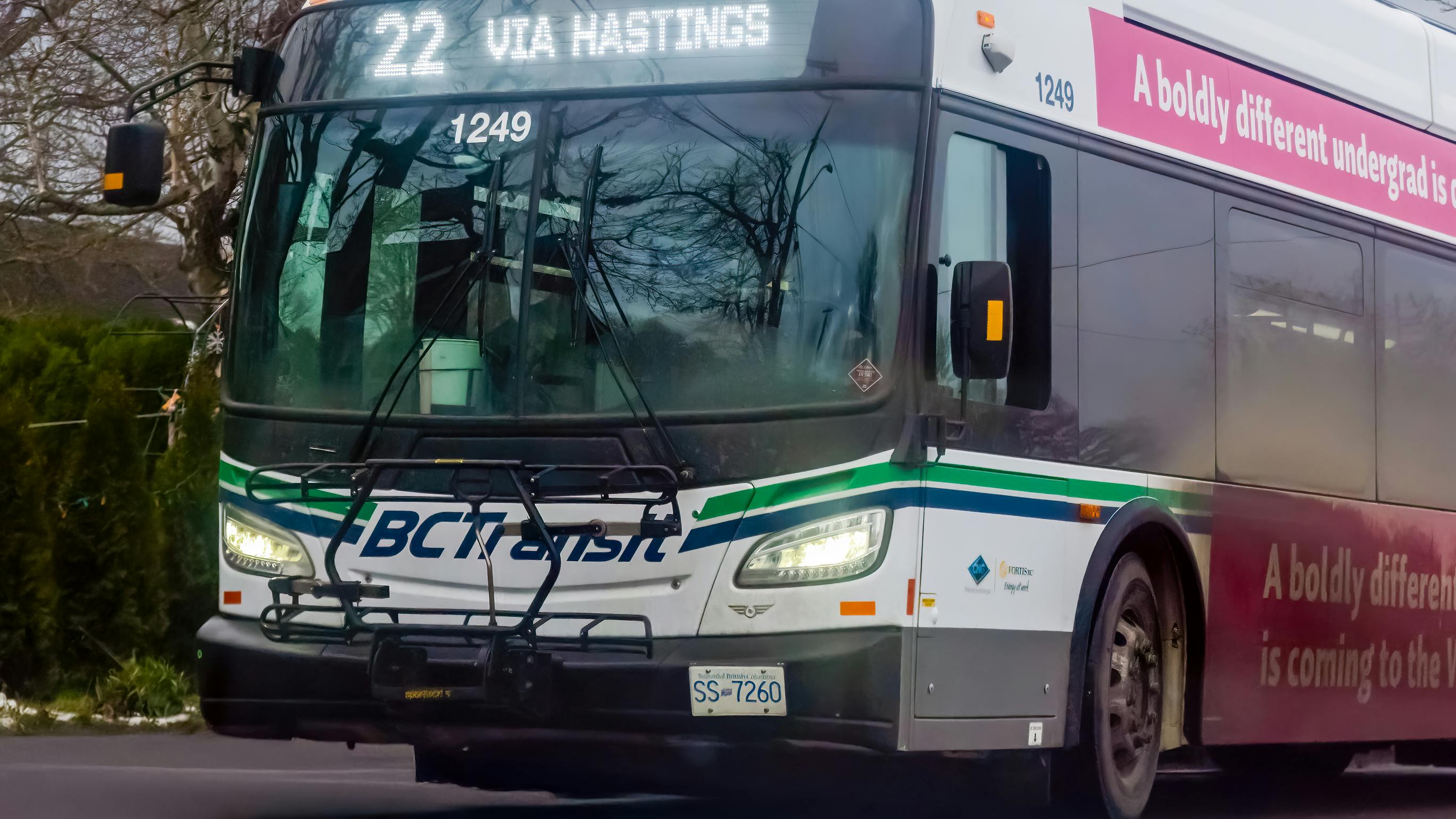 BC Transit CNG bus number 1249 displaying Route 22 Via Hastings on its destination board travels along a tree-lined road in Victoria, B.C., with a front-mounted bicycle rack and a pink university advertisement wrap visible on the side.