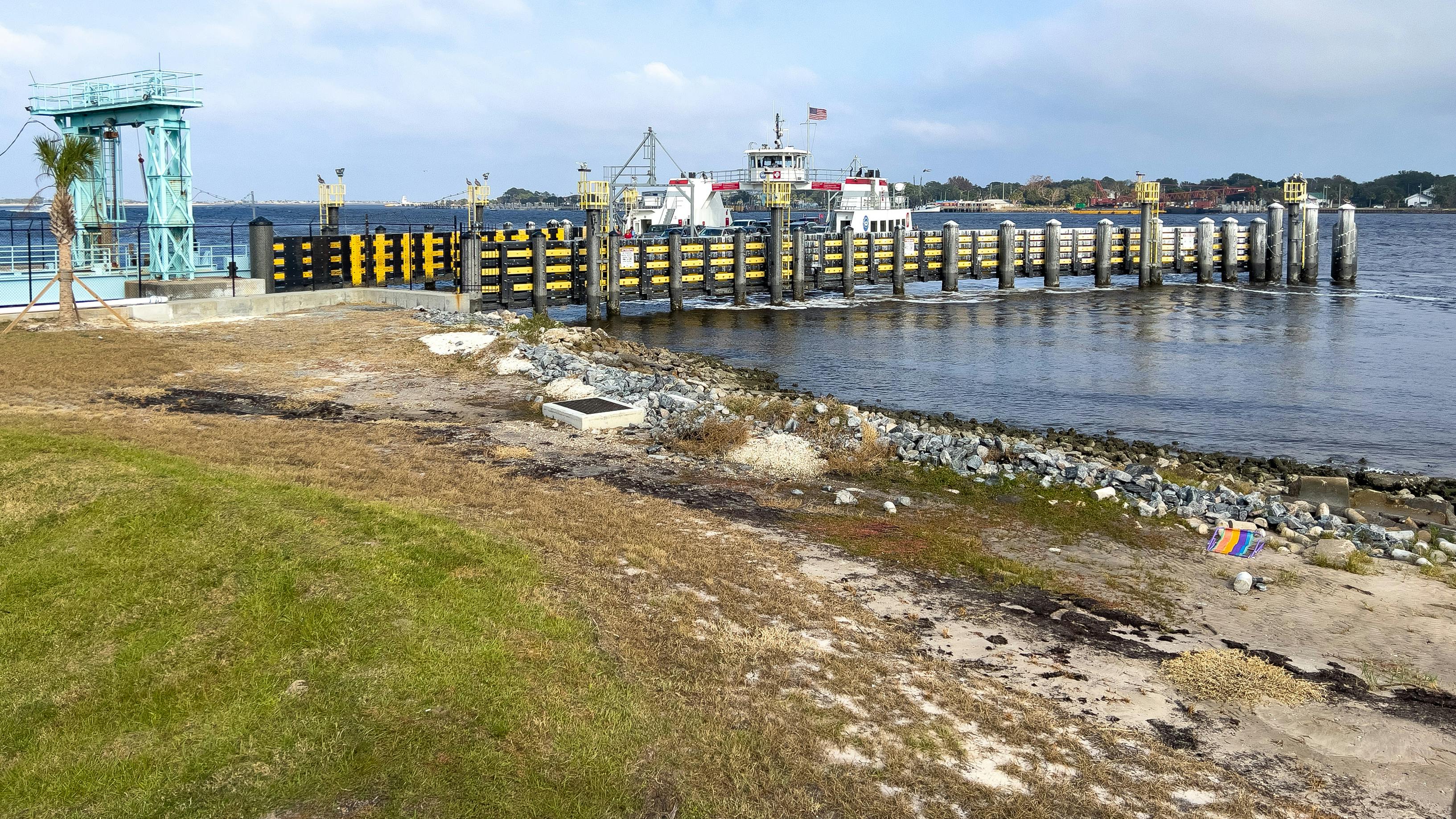 A ferry terminal dock with a moored ferry displaying an American flag, framed by a row of black-and-yellow fendered pilings and a teal loading crane, with a rocky shoreline and grassy waterfront in the foreground.