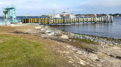 A ferry terminal dock with a moored ferry displaying an American flag, framed by a row of black-and-yellow fendered pilings and a teal loading crane, with a rocky shoreline and grassy waterfront in the foreground.