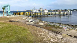 A ferry terminal dock with a moored ferry displaying an American flag, framed by a row of black-and-yellow fendered pilings and a teal loading crane, with a rocky shoreline and grassy waterfront in the foreground.