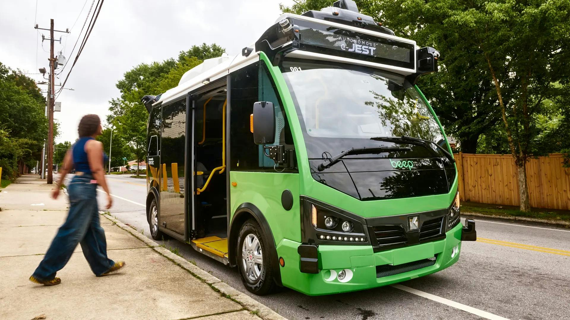 A green Beep autonomous 'e-JEST' shuttle bus, equipped with roof-mounted sensors and a wheelchair ramp, parked curbside on a tree-lined street as a pedestrian walks past on the sidewalk.