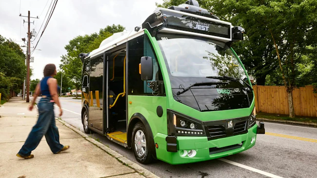A green Beep autonomous 'e-JEST' shuttle bus, equipped with roof-mounted sensors and a wheelchair ramp, parked curbside on a tree-lined street as a pedestrian walks past on the sidewalk.