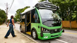 A green Beep autonomous 'e-JEST' shuttle bus, equipped with roof-mounted sensors and a wheelchair ramp, parked curbside on a tree-lined street as a pedestrian walks past on the sidewalk.