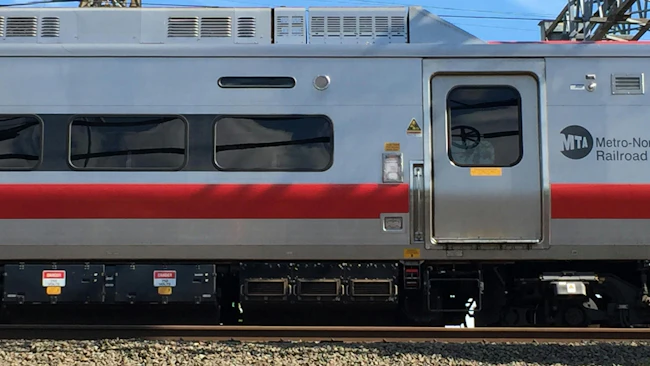 Close-up side view of an MTA Metro-North Railroad train car with its silver, red, and gray livery, showing the entry door, undercarriage equipment, and overhead catenary wires, with a stone platform edge in the foreground.