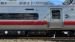 Close-up side view of an MTA Metro-North Railroad train car with its silver, red, and gray livery, showing the entry door, undercarriage equipment, and overhead catenary wires, with a stone platform edge in the foreground.