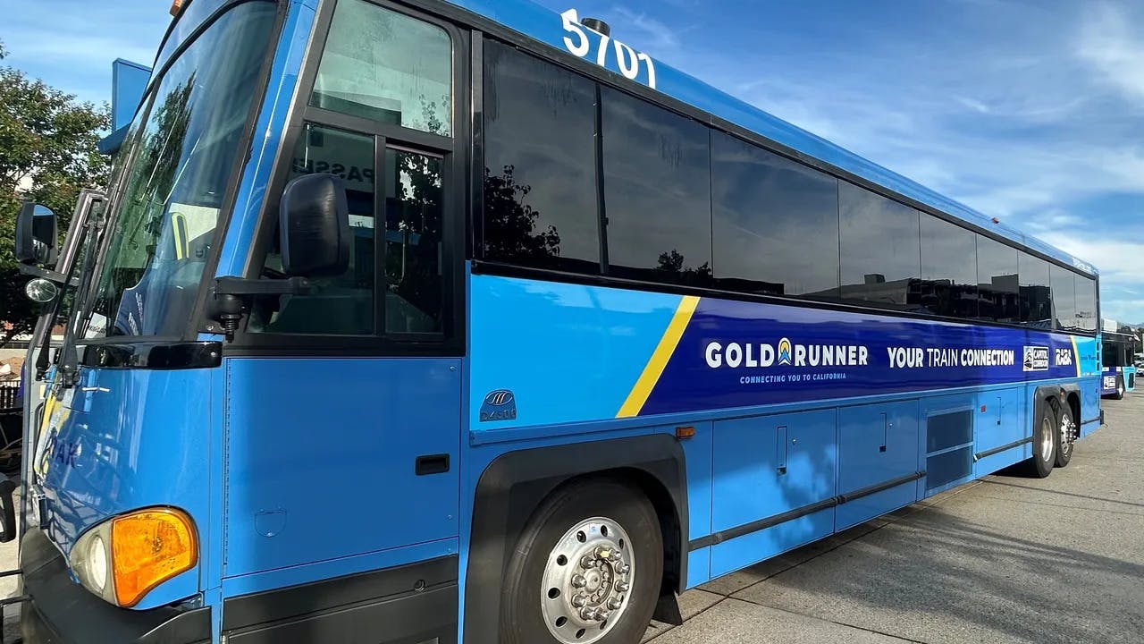 Gold Runner motorcoach bus number 5701 in a bright blue livery with a navy and yellow stripe displaying 'Gold Runner &mdash; Your Train Connection &mdash; Connecting You to California' branding along with Capitol Corridor and RARA logos, parked in a transit lot under a partly cloudy sky.