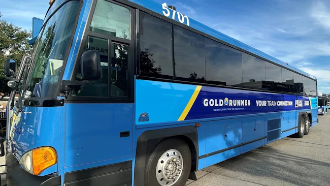 Gold Runner motorcoach bus number 5701 in a bright blue livery with a navy and yellow stripe displaying 'Gold Runner &mdash; Your Train Connection &mdash; Connecting You to California' branding along with Capitol Corridor and RARA logos, parked in a transit lot under a partly cloudy sky.