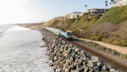 Aerial view of an Amtrak Pacific Surfliner train, locomotive 915, running along a rock-armored coastal rail line beside the ocean, with cliffside homes and palm trees above in San Clemente.