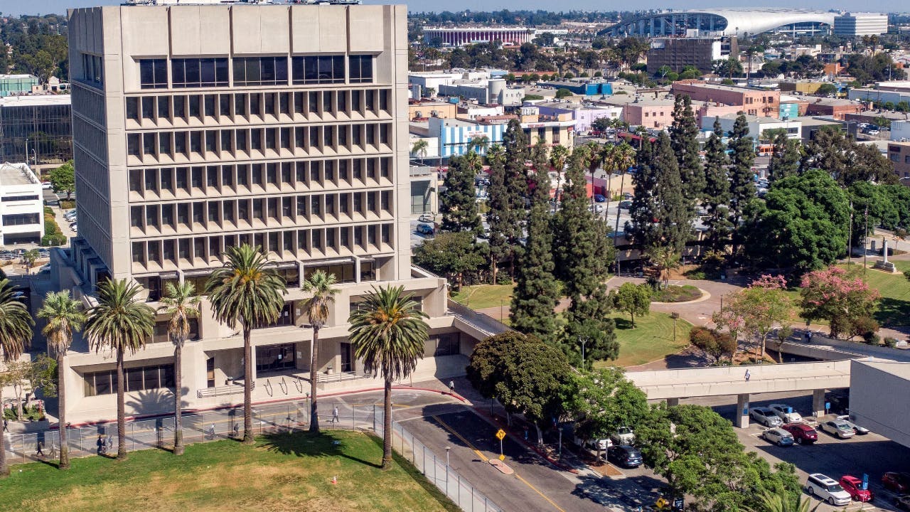 An aerial view of a mid-century concrete office tower in Downtown Inglewood, Calif., surrounded by palm trees and green space, with the broader Inglewood urban landscape, retail corridors, and a large stadium structure visible in the background.